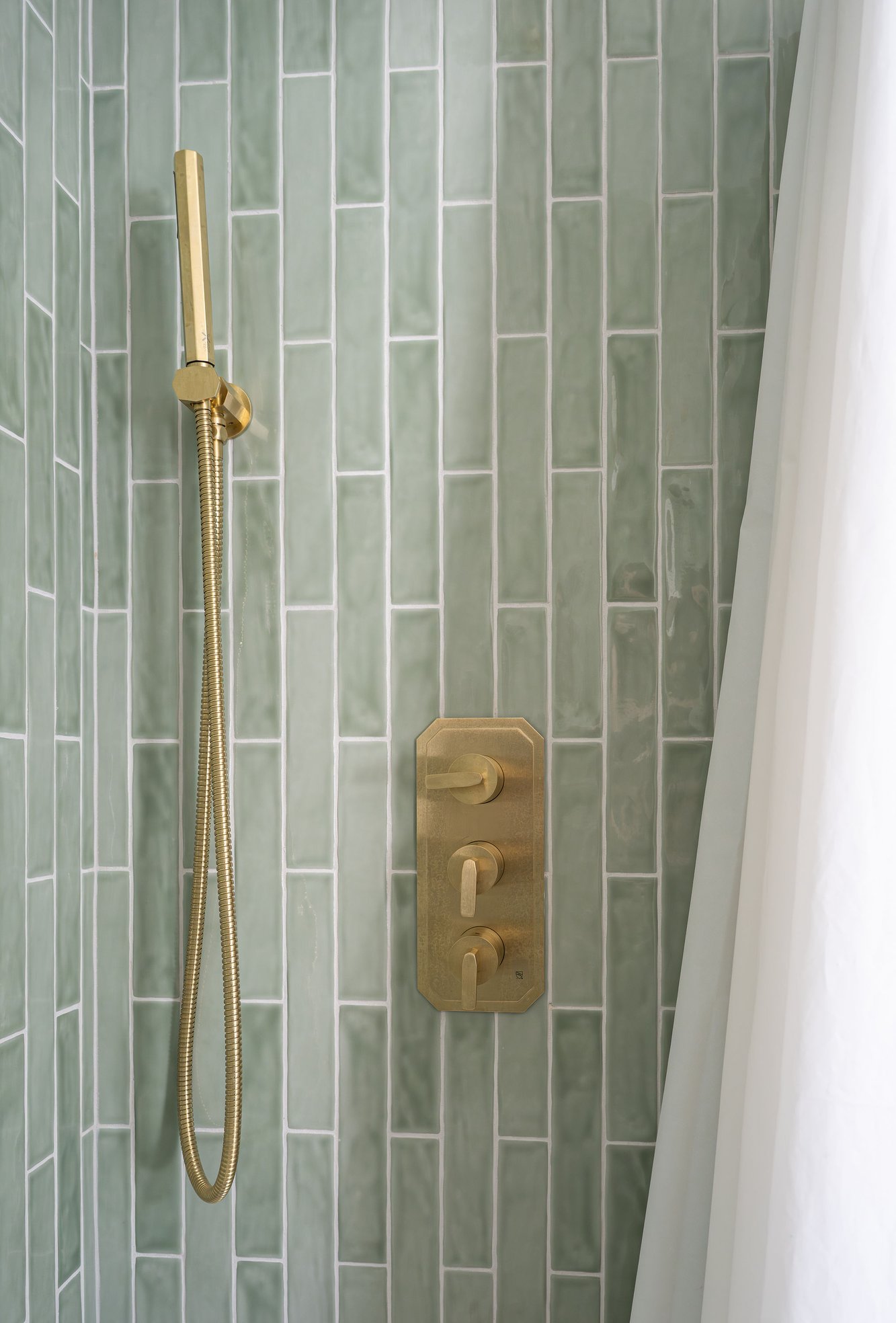 Brass handheld shower and controls installed against glossy sage-green vertical tile. Brass handheld shower and controls installed against glossy sage-green vertical tile.