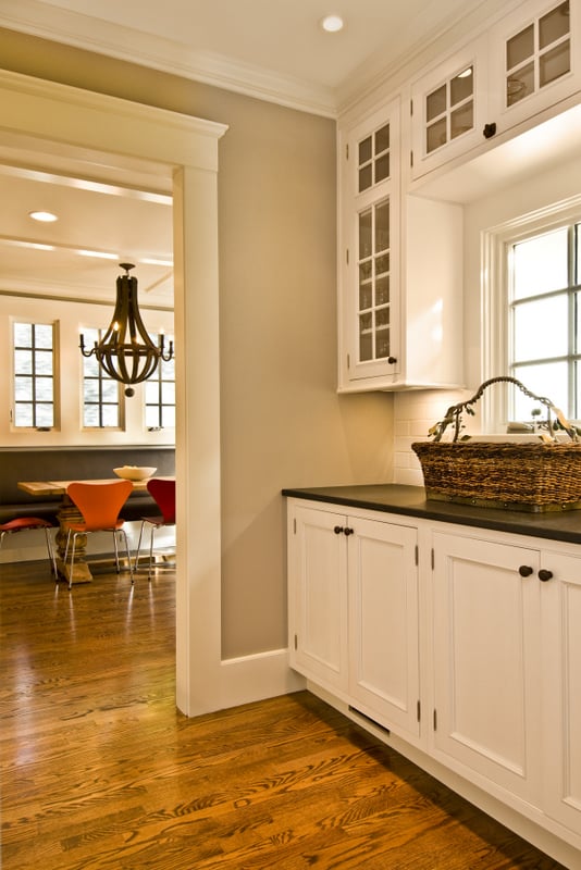 Renovated butler's pantry with white shaker cabinets, black countertop, and glass-front upper cabinets, opening into a dining room with a wood chandelier and orange dining chairs.