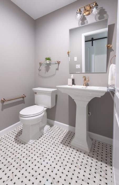 Front view of a remodeled bathroom in a home in Essex County, NJ, with brass fixture, a handlebar by the toilet, a porcelain faucet, and checkered pattern tile