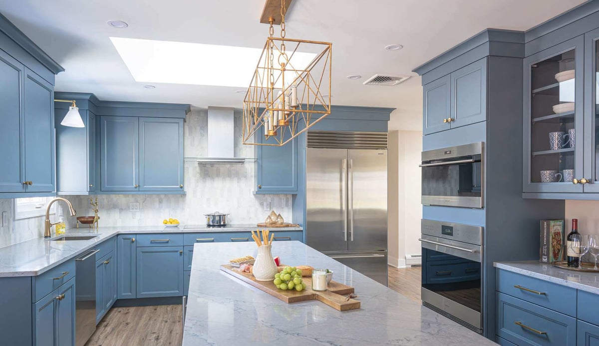 A full kitchen perspective emphasizing the cohesive design elements including stainless steel appliances, blue cabinetry, marble surfaces, and tasteful decorative touches. In Montclair, NJ | G&amp;L and Sons Renovations.
