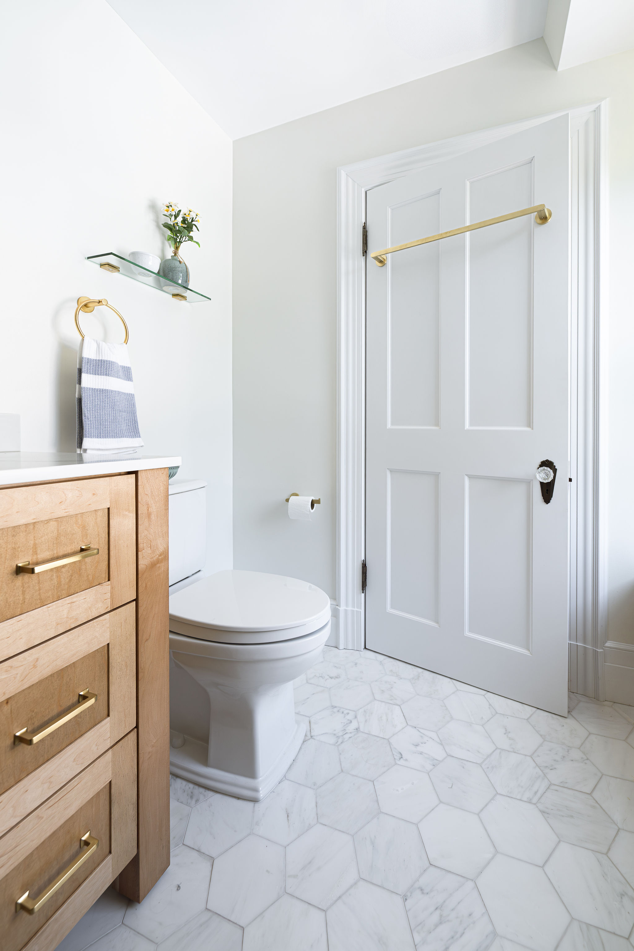 Bright remodeled bathroom with a natural wood vanity, brass hardware, white toilet, and marble hexagon floor tile.