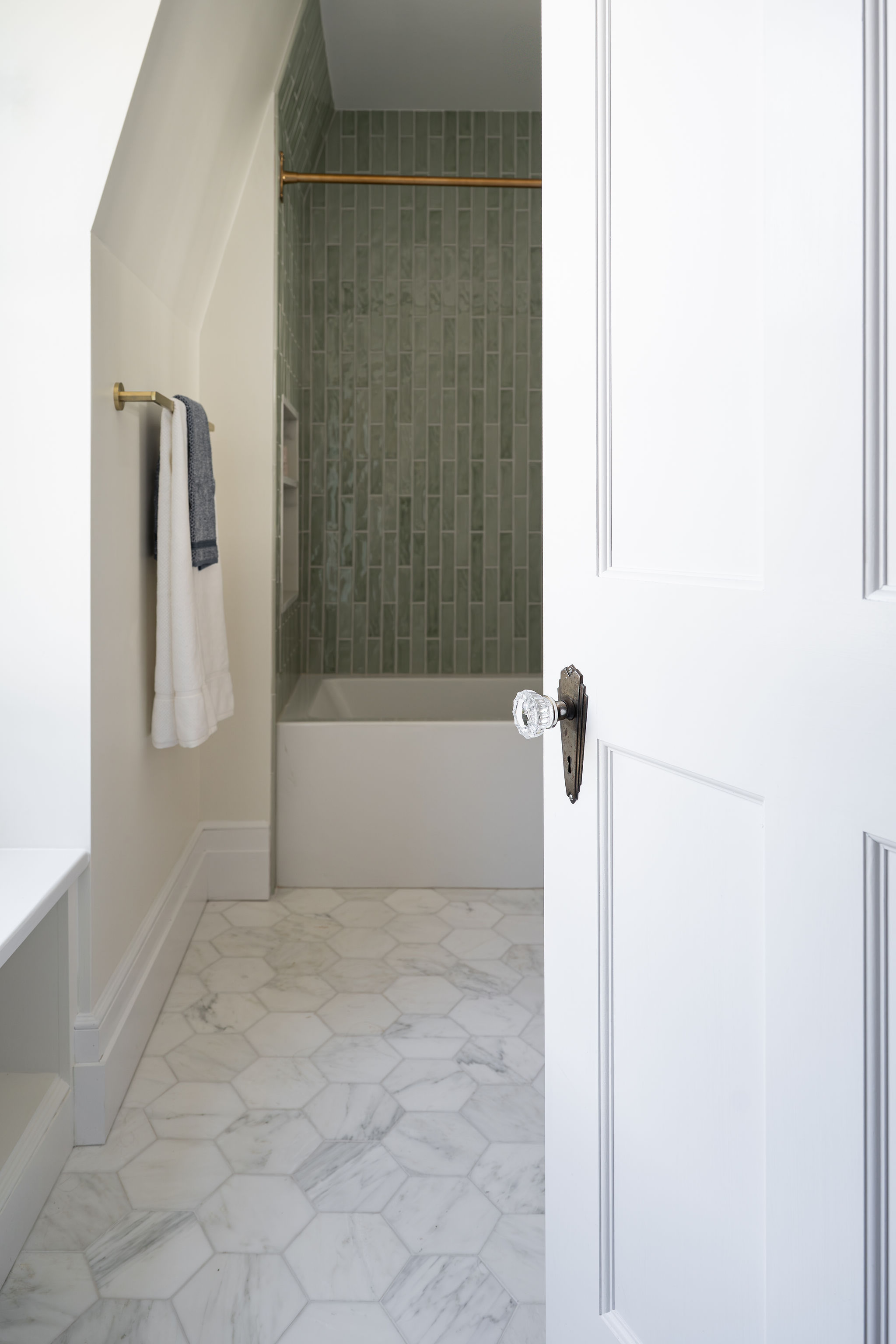 View through a white paneled door into a remodeled bathroom with green shower tile and marble hexagon flooring.