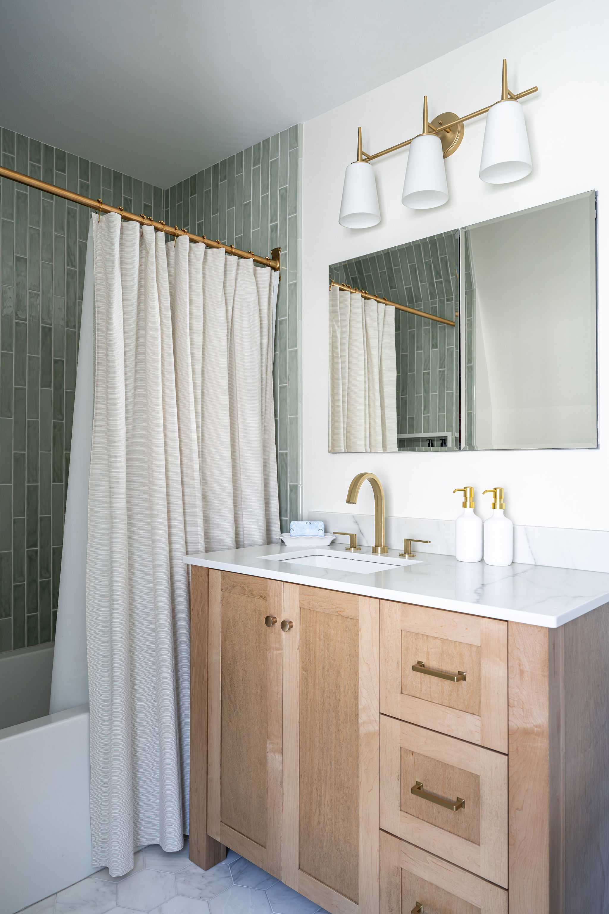Bathroom vanity with natural wood cabinetry, marble countertop, brass fixtures, and green tiled tub surround.